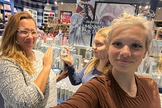 A woman and two teenaged girls are standing in a store that sells fragrances and beauty products. The woman on the left is making a funny or disapproving face while raising her hand toward a spray bottle held by one girl in the middle, as if gesturing “no.” The girl on the right, smiling at the camera, holds the phone for a selfie. The store’s background features various product displays, including a sign reading “inspired by Mesmerizing Metallics.”