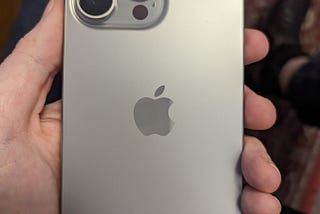 The back of an iPhone 15 Pro Max in Natural Titanium, being held in a white hand.