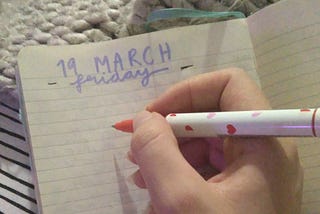 My hand holds a pen on top of my journal page. My journal page has the heading 19th March, Friday, on it.