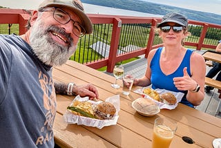 The author and his wife sit at an outdoor brewery table overlooking Seneca Lake, with sandwiches, chips, and drinks in front of them. The lake and wooded shoreline stretch behind them under a bright sky.