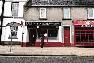 Dilapidated, 2 or 3-story stone buildings with blank windows — one houses a fish & chip shop called ‘Salt and Vinegar’, another a Chinese takeout. An indistinct person walks along the pavement in front. At the bottom of the picture, bright yellow double-lines edge the tarmac road and underline the curb.