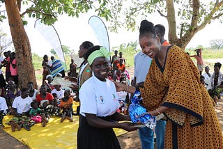 A young black girl wearing a white t-shirt with the UN Trust Fund logo, a green headband and black skirt, is seen smiling receiving an award from a young black women wearing a yellow and black dress. In the background we can see people sitting and standing outside.