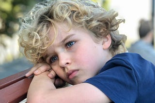 A young boy with curly blond hair and a blue shirt rests his head on his arms, looking worried and deep in thought. His expression is tense and distant, with furrowed brows and slightly pursed lips.