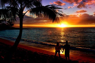 Beautiful brightly colored orange, yellow, purple, blue ocean beach sunset with black silhouettes of a palm tree and a couple (man & woman) holding hands in the suns reflection on the wet beach sand.