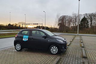 Photo of a Peugeot 108 parked in a parking lot next to a motorway. It has a sticker on it that says Juuve, the name of the rental company.