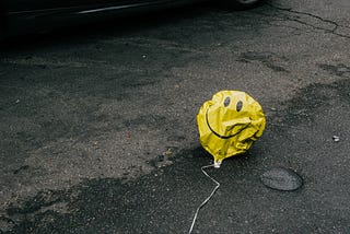 Picture of a deflated, yellow smiley-face balloon that’s on a road being run over by cars.