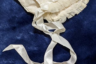 A satin off-white baby bonnet with lace trimming and two lace ribbons streaming beneath it. The bonnet is on top of a blue fake fur surface for better contrast.
