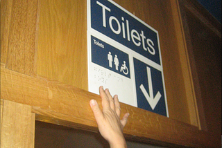 Picture of a braille sign above a door. A hand is reaching up, but the braille is too high to be felt.