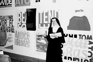 Photography of Sister Corita Kent stands in front of her work, including for Eleanor, at Immaculate Heart College in 1964.