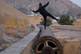 The back of a person in black clothing, and black hat, balancing on one leg on a large pipe while facing a mountain in the distance.