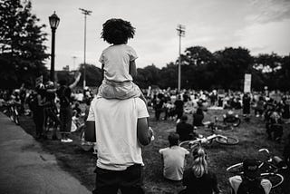 Black and white photo of a young child sitting the shoulders of an adult overlooking a green space populated with sitting people.