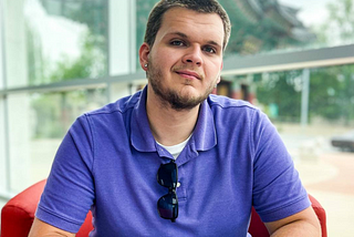 A photo of Isaac Mashman in a purple polo shirt, with sunglasses in his collar, smiling and looking at the camera.