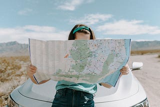 Woman sitting on car bonnet looking at map held in both hands hiding her face