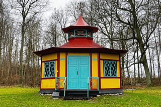 A small, hexagonal, 18th-century teahouse, painted bright yellow. with red and turquoise trim. Surrounded by bare wintery trees and set in a bright moss-green lawn.