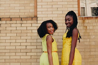 Two young black girls in yellow dresses, smiling, looking toward the photographer, standing in front of a brick wall.