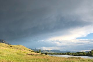 a picture of a storm rolling in over a mountain and sun-kissed wild grass hills next to a river in Paradise Valley, Montana