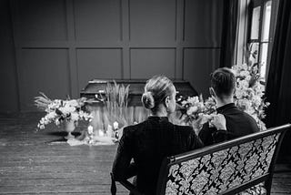 woman and man dressed in black with their backs toward the camera, sitting on a bench facing a casket surrounded by flowers and candles