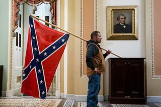 An often-republished photo of a January 6 rioter standing in the Capitol Building, holding a large confederate flag.