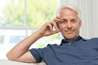 Older attractive white man with gray hair and a blue shirt staring into camera with a closed mouth smile, and leaning on his right arm.