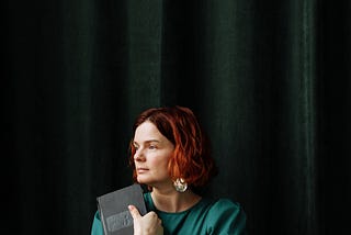 Woman with red hair holds a closed book and looks out of a window
