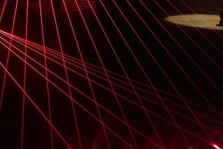 Red laser beams criss-cross the darkness. In the top right corner, a person stands in a pool of light.