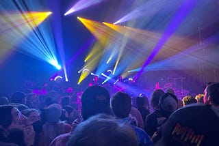 A crowd of music lovers watching the band Spafford on stage at the Park West venue in Chicago. There are bright blue, yellow, and purple lights shining out onto the audience.