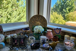 Knick knacks including an owl mug with a hydrangea blossom in front of two windows on a kitchen ledge.