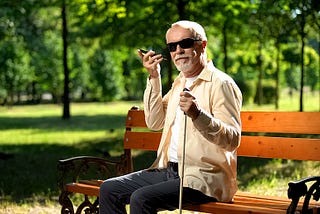 Older man wearing dark glasses and holding a white cane in left hand, sitting on park bench holding smartphone to his ear with right hand.