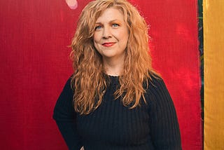 Photo: Mindy Tucker. Woman with long red hair, wearing a navy sweater and navy pants. The early-autumn sun shines through trees visible above the red and gold backdrop.