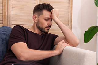 A man with short brown hair and a beard sits on a couch, resting his arm on the armrest. He appears to be deep in thought, with his hand resting on his forehead and his gaze focused downward. He is wearing a dark t-shirt, and a wooden panel background is partially visible behind him.