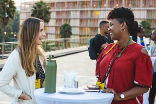 A white woman and a black woman smile and talk together over a cocktail table