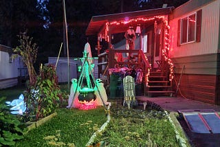 A porch is decorated with glowing orange lights, spooky figures, and a gravestone for Halloween. In the yard, a cauldron surrounded by eerie decorations emits a mysterious green glow.