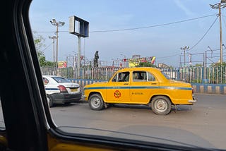 A view through a car window showing a classic yellow taxi in Kolkata, India. The taxi is parked next to a white car, with a fence and street signs visible in the background. The sky is clear, and the streets appear calm.