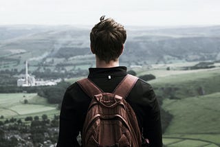 Male with short dark hair facing a countryside in front of him. His back his turned and he he wearing a brown backpack.