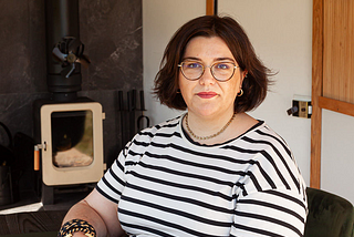 A lady with short dark brown hair wearing glasses and a striped T shirt sits in front of a log-burning stove