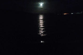 Photo of the moon coming out from the clouds and shining over a bay —  leaving a trail of light on the dark water.