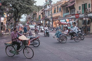 A photo of traffic in Hanoi, with various modes of transportation shown, including motorbikes, a bicycle, and a rickshaw