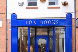 Photograph of the front of the Fox Books bookshop, shows a blue front, with door and windows either side. Red and orange bricks on either side, then a grey roof above. Pavement in front, with a wooden foldable sign