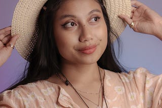 A portrait of myself wearing a pink floral dress, both hands reaching up touching the brim of my straw hat and looking to the right side with a smile.