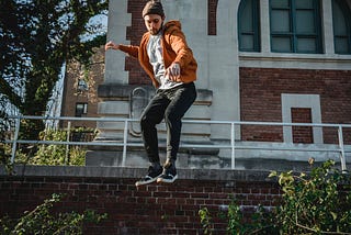 A young man wearing an orange hoodie and toque jumps off a brick wall in a parkour move.