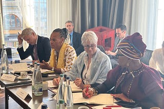 Four people at a meeting table, with papers and water bottles. Behind them are two additional people sitting in the back. They are in a corner conference room.