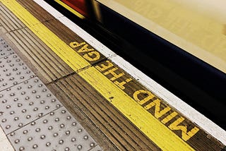 Image of a subway platform with a yellow tactile strip that reads ‘MIND THE GAP’ in bold letters. The platform has a textured surface with raised dots for safety. A blurred train is visible in the background.