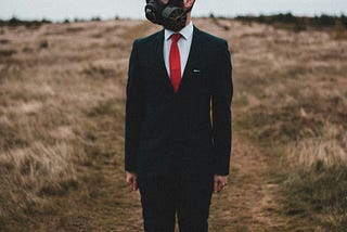 White male (man) with combination top-patterned baldness and short dark side and occipital hair, with head slightly askew to the right, arms at his side, hands clenched with thumbs tucked, standing in dry grassland wearing a gas mask, black tailored suit with a button-up white collar shirt with a red tie and brown oxford loafers.