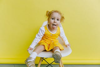 Little girl with her hair tied up, sticking her tongue out, she is wearing a yellow dress and the background is yellow, Jessie Waddell, Medium