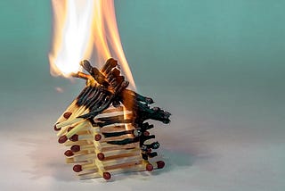 Small house made of matchsticks partially consumed by flames, on a white to aqua gradient background.