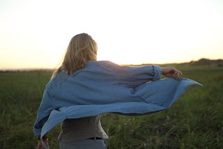 Woman with her shirt billowing in the breeze walks in a field after sunset