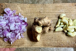 On a wooden tray, chopped onions, ginger, and peeled garlic are placed in individual groups.