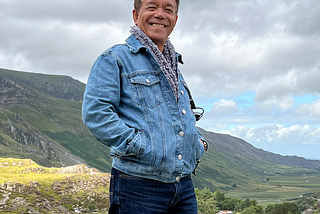 My father wearing a denim jacket and a scarf with Welsh mountains and a cloudy sky as the backdrop.