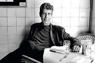 A black & white photo of Anthony Bourdain sitting at a restaurant table.