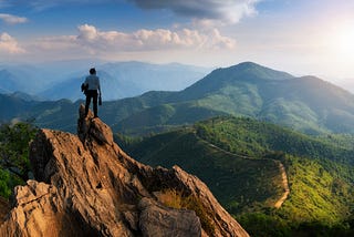 A businessman with a briefcase stands on a mountain peak overlooking more mountains as far as the eye can see